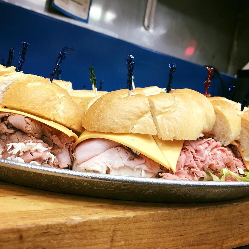 Tray of mini sandwich sliders with toothpicks, filled with turkey, ham, and cheese on a wooden table.