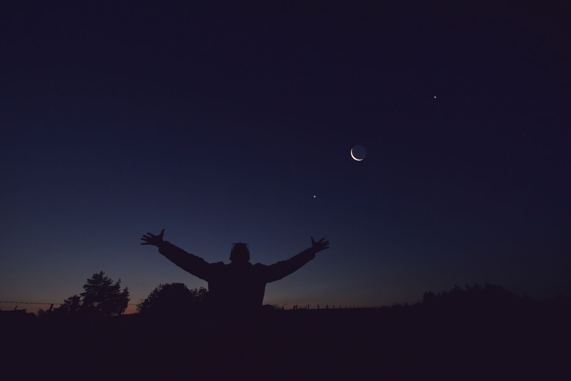 Silhouette of a person with arms raised under a crescent moon in a twilight sky