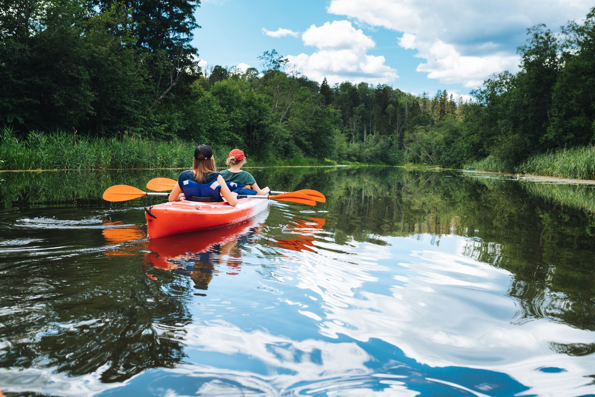 Two people paddling a red kayak on a calm river surrounded by green trees and blue sky