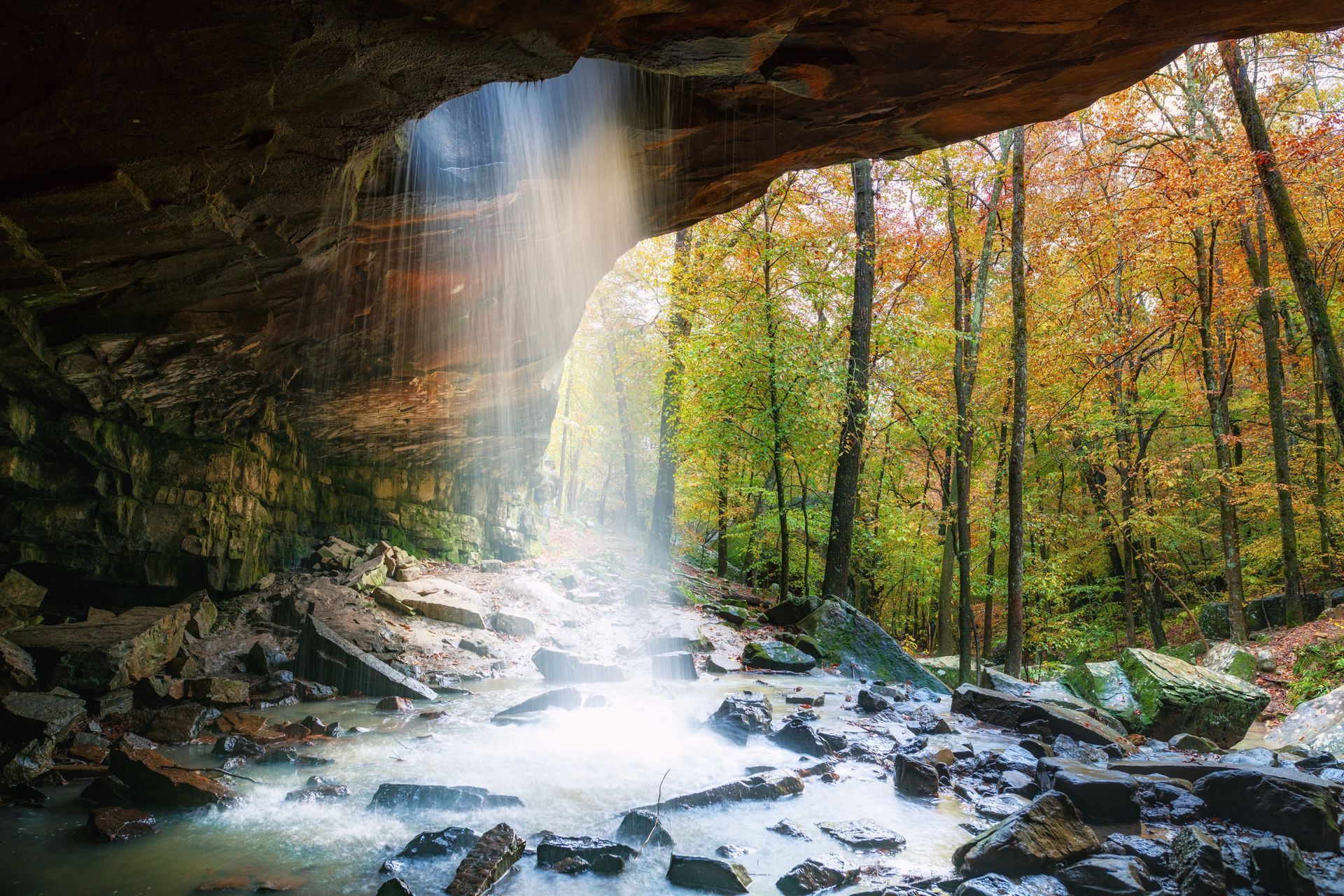 Waterfall spilling from a rocky overhang into a stream, framed by autumn trees and mossy rocks.