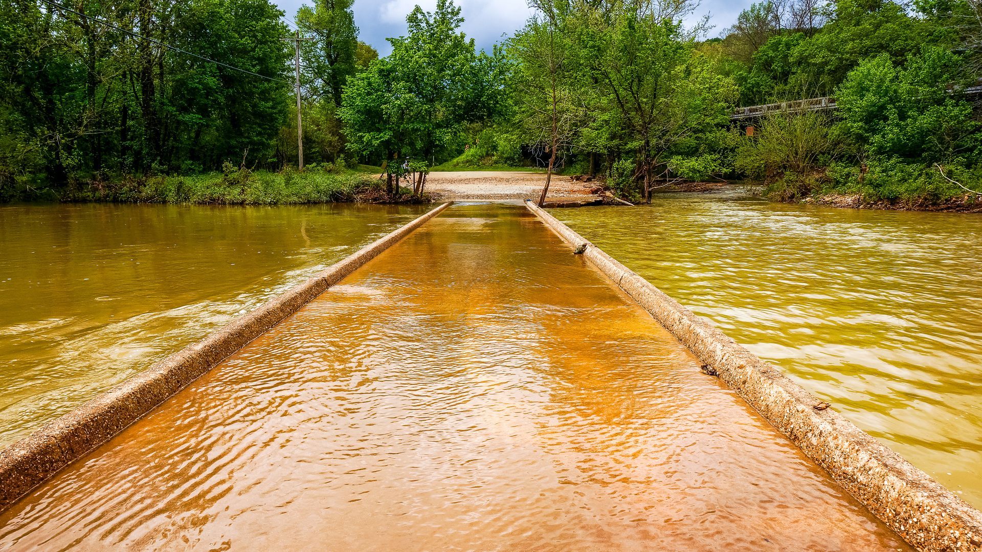 Flooded dirt road with muddy water leading toward green trees under a cloudy sky