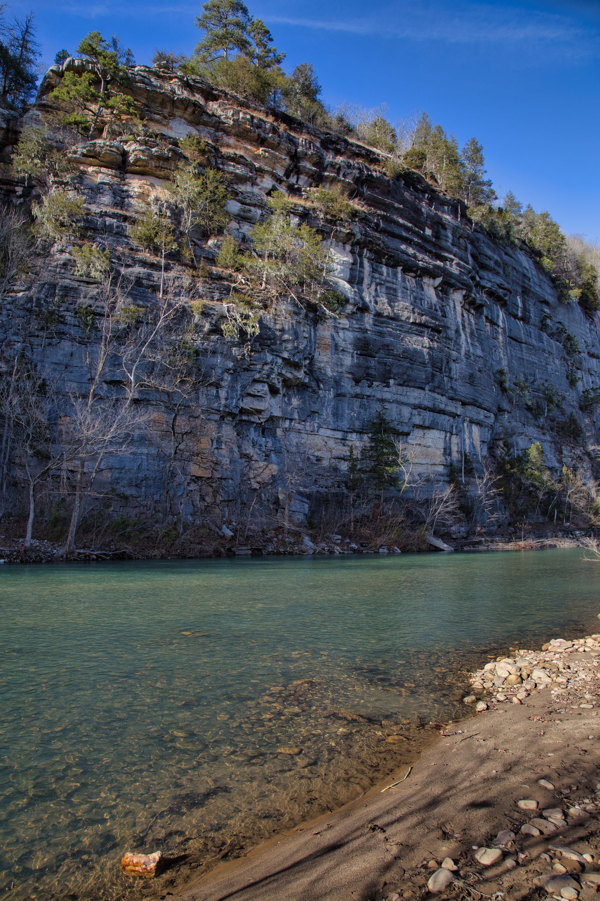Steep rocky cliff beside clear turquoise river under a blue sky, with a pebbled shore in the foreground