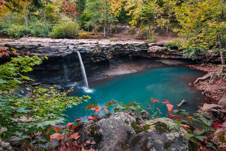 Waterfall spilling into a turquoise pool in a rocky forest gorge with autumn foliage