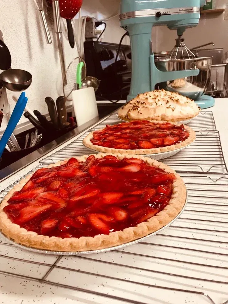Two strawberry pies cooling on a wire rack in a kitchen, with a mixer and utensils nearby.