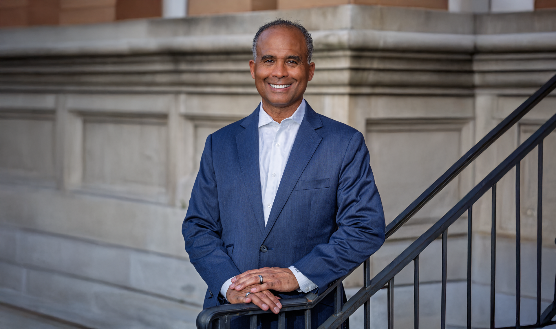 Smiling man in a blue blazer standing on outdoor stone steps beside a black railing