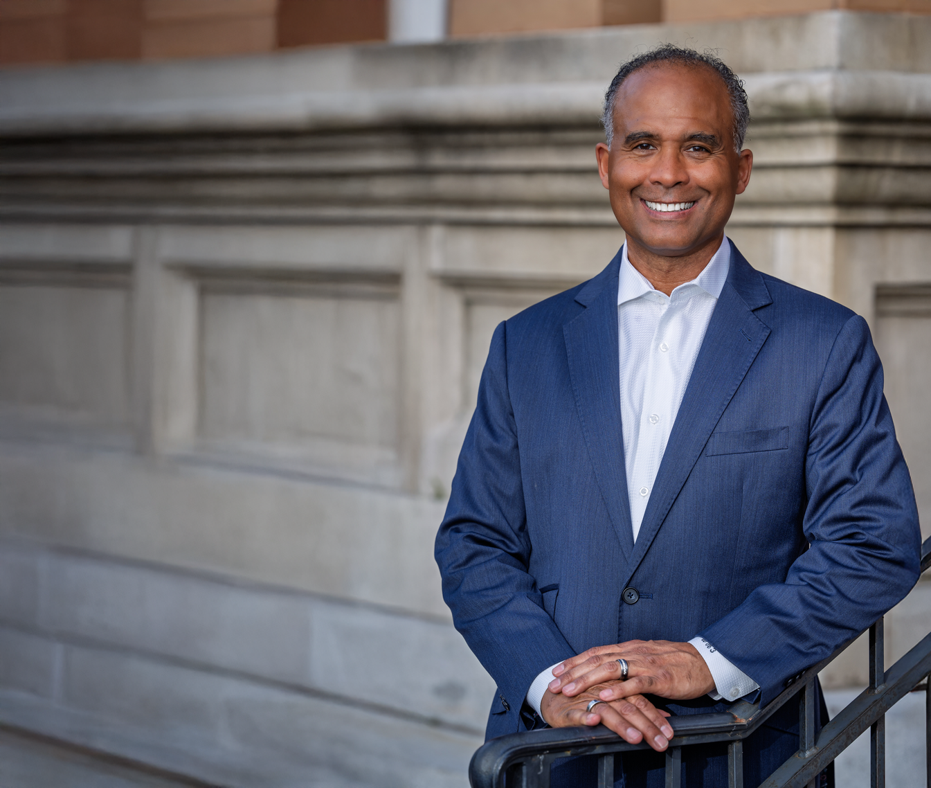 Smiling person in a blue suit standing by a stone railing outdoors