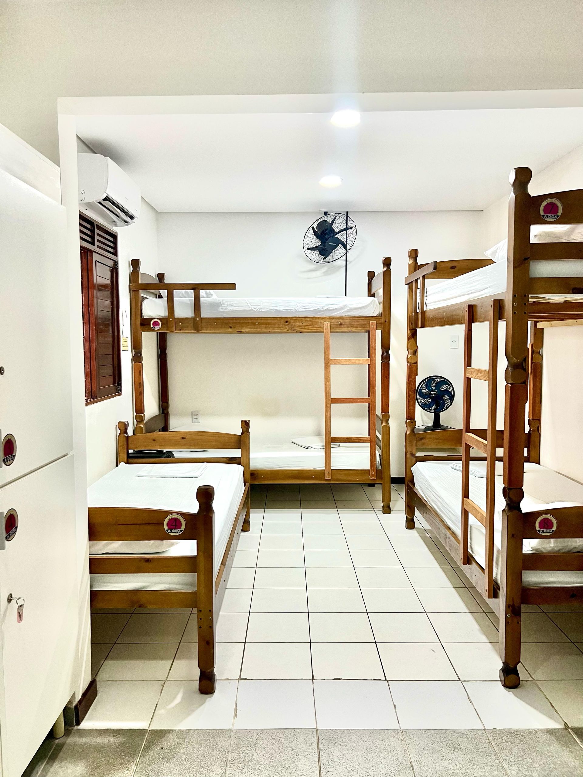 Quarto de dormitório com três beliches de madeira, piso de azulejo branco e iluminação forte no teto.