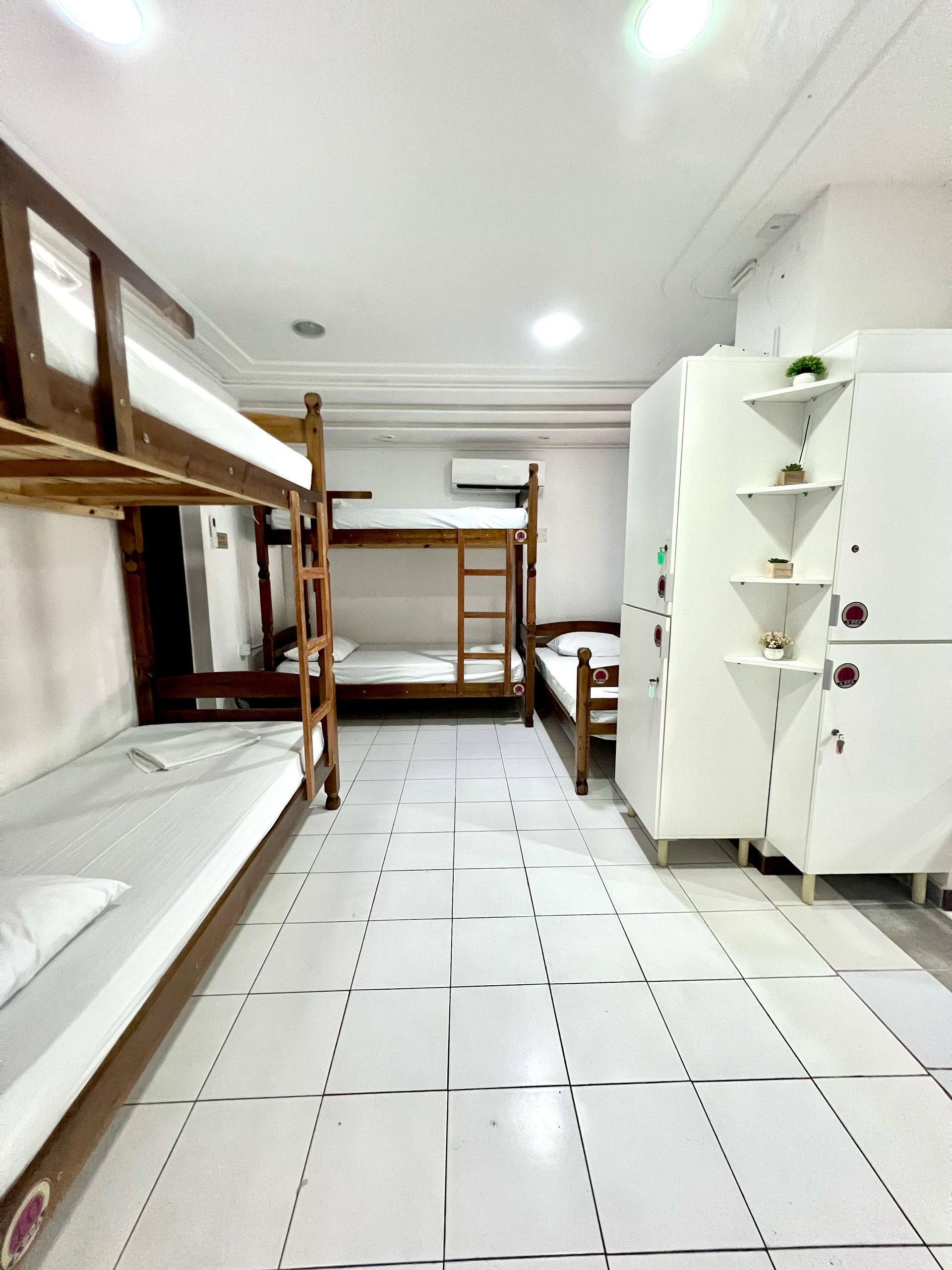 Quarto de dormitório comprido e estreito com beliches, piso de azulejo branco e armários brancos para guardar roupas.