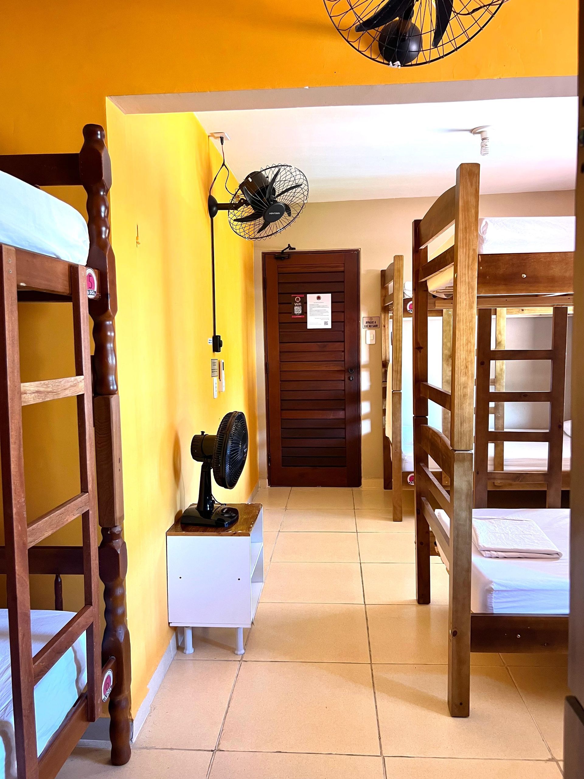 Quarto de dormitório com beliches de madeira, piso de azulejo, paredes amarelas e uma porta no fundo.