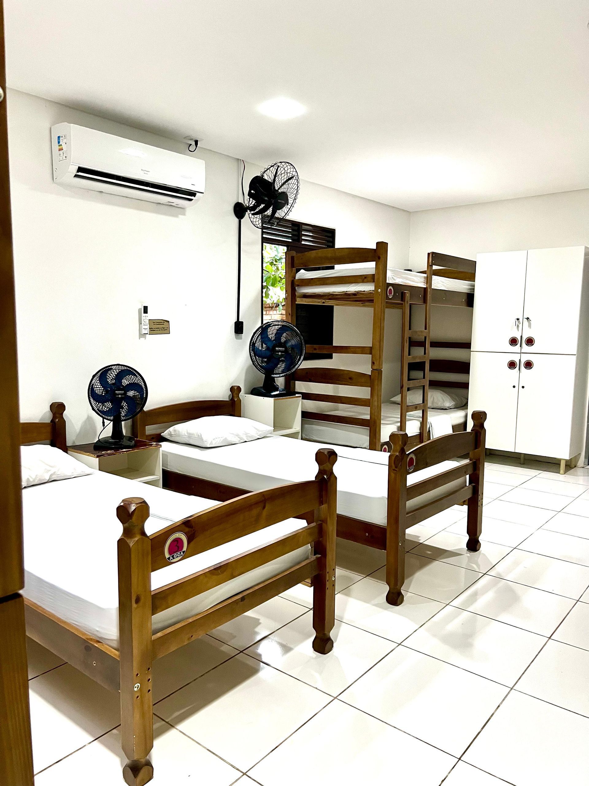 Quarto de dormitório com duas camas de beliche de madeira, ventiladores, ar condicionado e piso de azulejo branco.