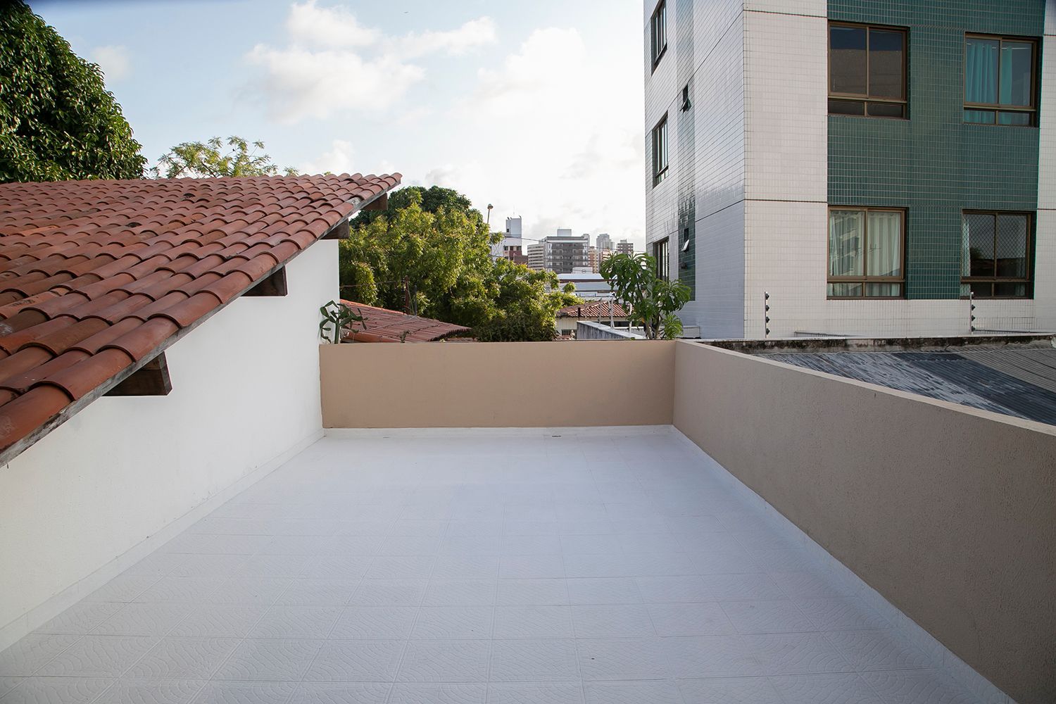Terraço vazio na cobertura com piso branco, telhado de telhas vermelhas à esquerda e prédios da cidade ao fundo.
