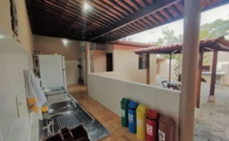 Cozinha externa coberta com pia, bancada e lixeiras coloridas ao lado de um pátio com piso de azulejo.