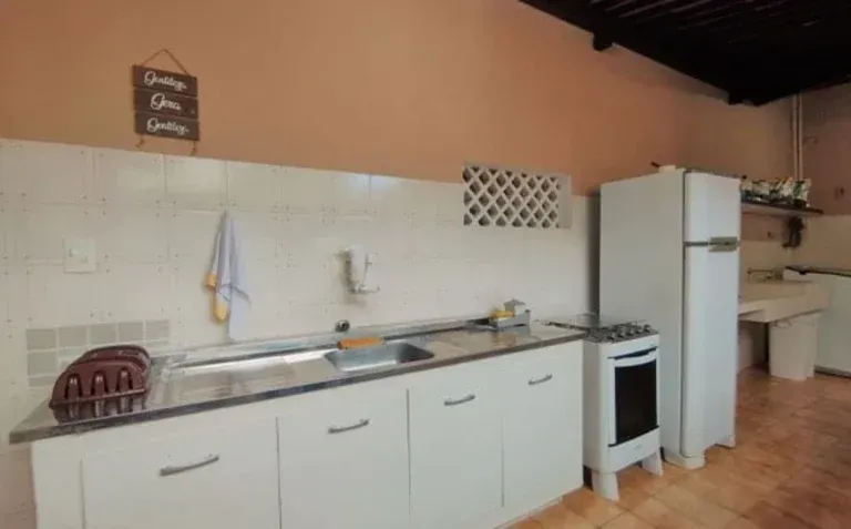 Cozinha externa espaçosa com armários brancos, pia, fogão e geladeira sob um pátio coberto.