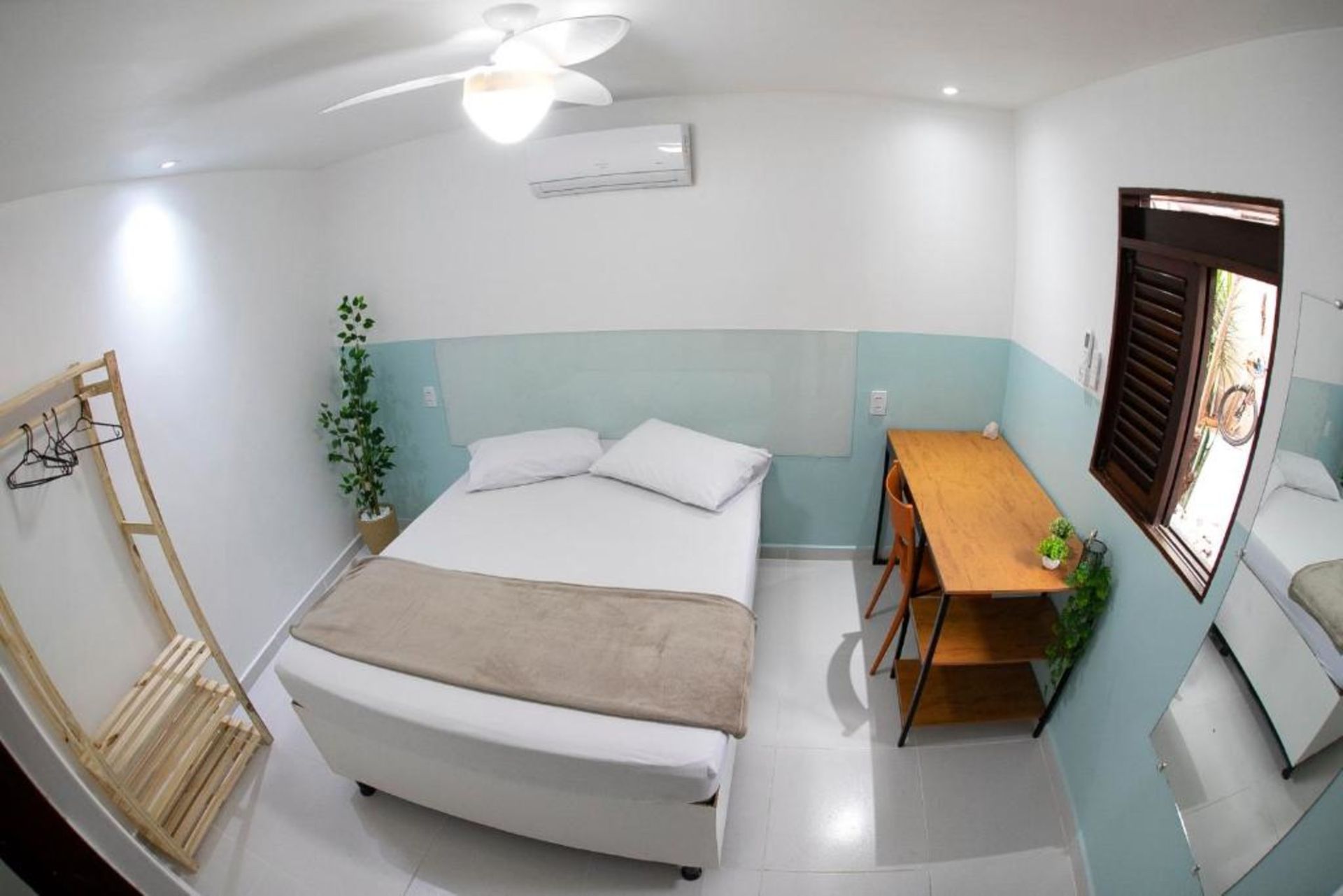 Quarto pequeno com cama, escrivaninha de madeira, persianas, ventilador de teto e paredes azul-claras.