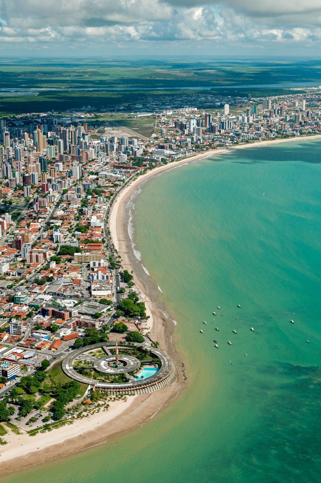 Vista aérea de uma praia de areia curva ao lado de águas turquesa e um denso horizonte urbano costeiro.