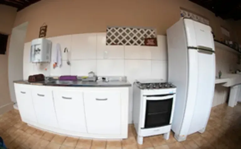 Cozinha pequena e branca com armários, fogão, pia e geladeira alta sobre piso de azulejo bege.