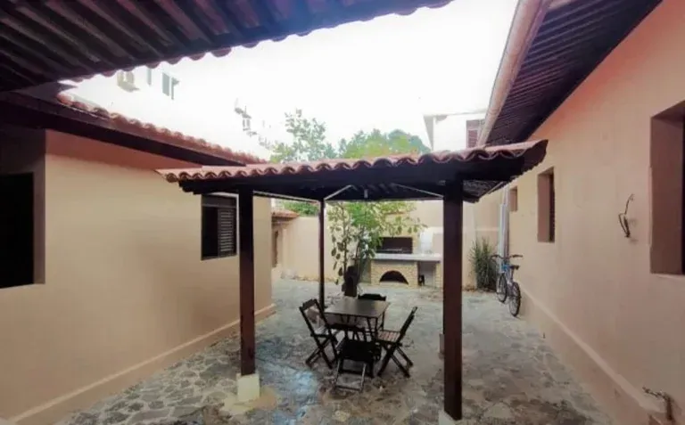Pátio interno com mesa e cadeiras sob uma pérgola entre dois edifícios cor de pêssego.