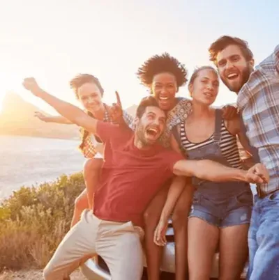 Amigos rindo e posando à beira-mar ao pôr do sol, com os braços erguidos em comemoração.