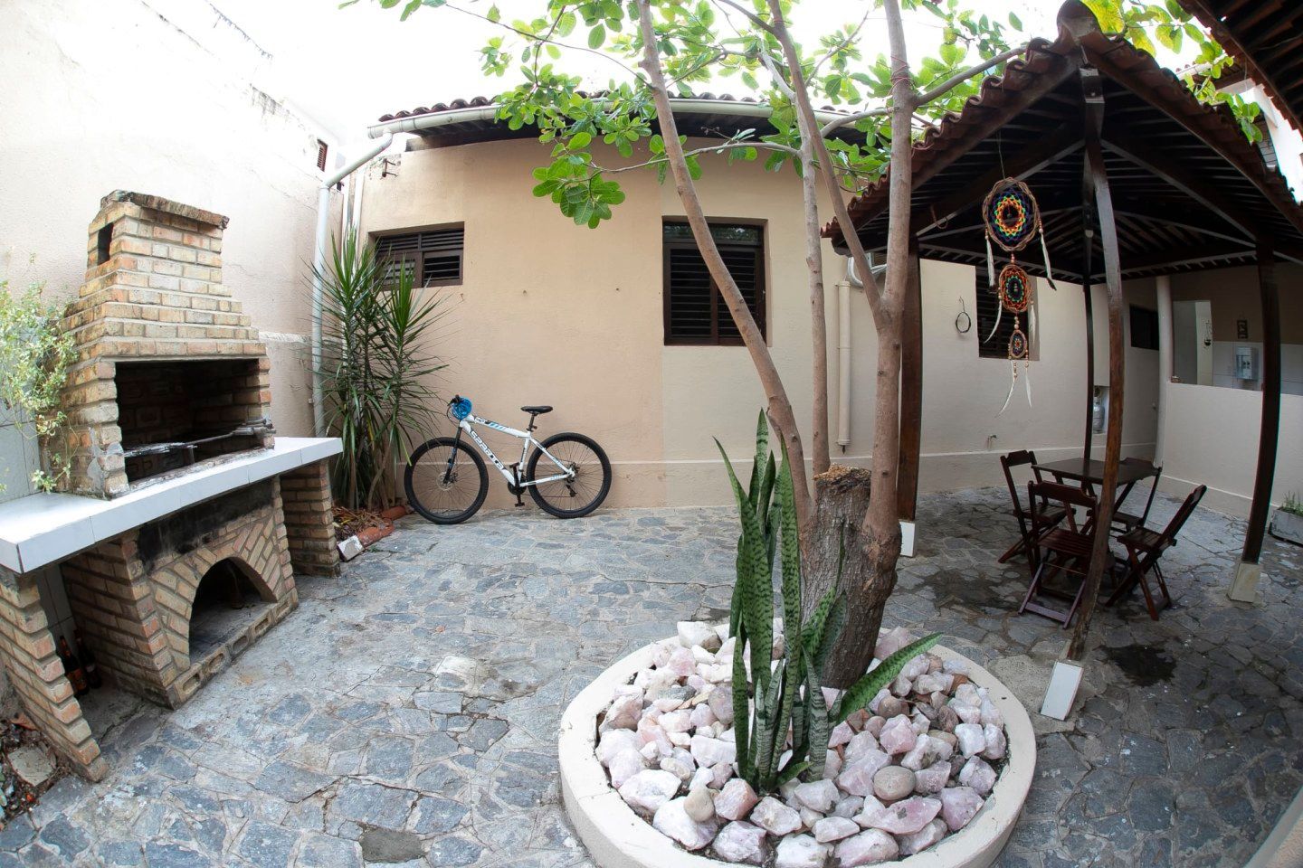 Pátio nos fundos com piso de pedra, churrasqueira de tijolos, cactos em vaso, bicicleta e área de estar sombreada.