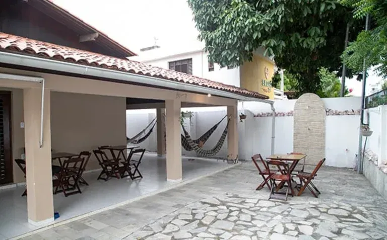 Pátio coberto com mesas e bancos de madeira em um pátio de pedra ao lado de um muro branco e árvores.