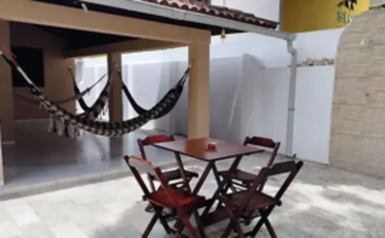 Pátio com mesa e cadeiras de madeira sob uma rede ao lado de um telhado de telhas e parede branca.