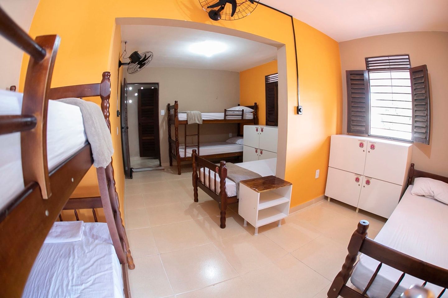 Pequeno quarto laranja com beliches, escrivaninha, cadeira, janela e caixas empilhadas no chão.