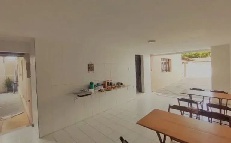 Área de jantar em um pátio luminoso e branco, com mesas e bancos de madeira, que se abre para um pátio interno.