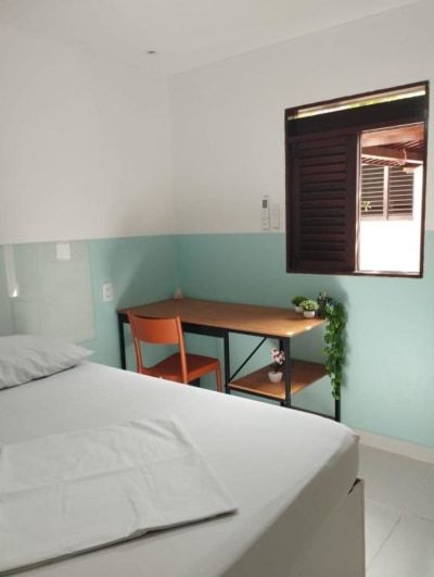 Quarto pequeno com cama arrumada, escrivaninha de madeira, paredes verde-menta e janela aberta com persianas.