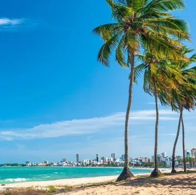 Praia tropical ensolarada com palmeiras, água turquesa e o horizonte distante da cidade sob um céu azul límpido.