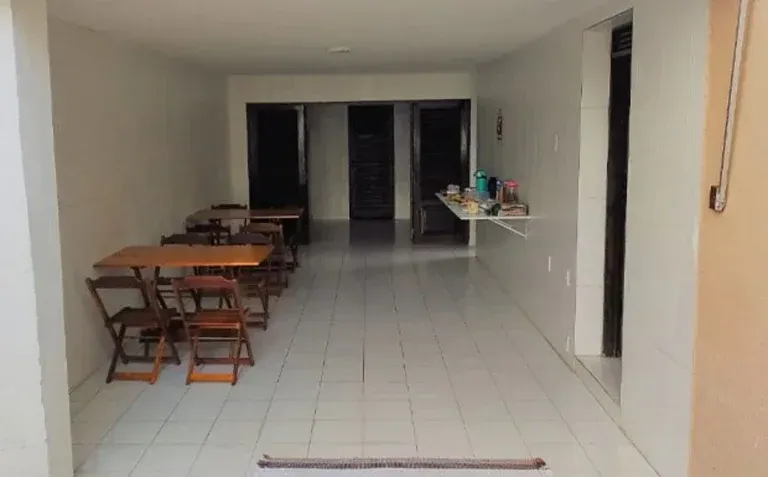Sala comprida com piso de azulejo, mesas de madeira à esquerda e um balcão no fundo.