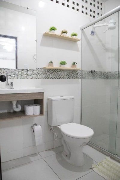 Banheiro pequeno e moderno com box de vidro, vaso sanitário, pia, espelho e prateleiras decorativas.