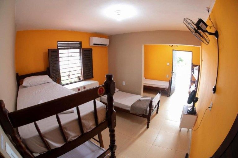 Quarto pequeno com duas camas, paredes laranja, persianas, ventilador de teto e porta aberta para um corredor iluminado.