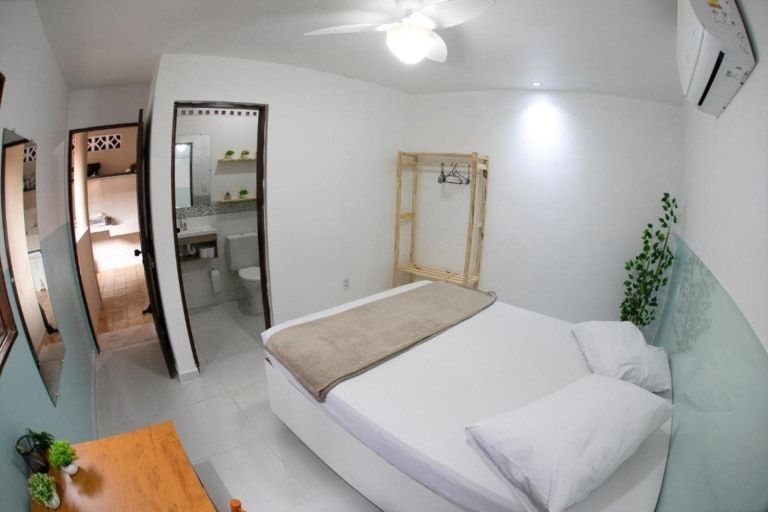 Quarto minimalista e luminoso com cama branca, cadeira de madeira, espelho, banheiro aberto e ar-condicionado de parede.