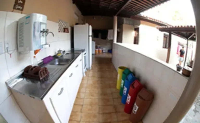 Cozinha estreita com armários brancos, piso de azulejo, bancada com pia e lixeiras coloridas ao longo de uma meia parede.