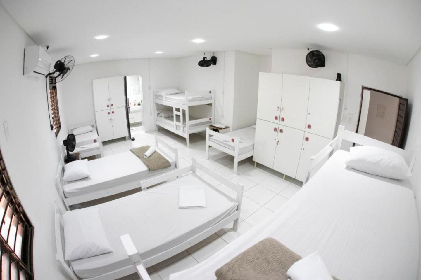 Quarto de dormitório claro e branco com beliches, camas de solteiro, guarda-roupas e uma pequena área de escritório.