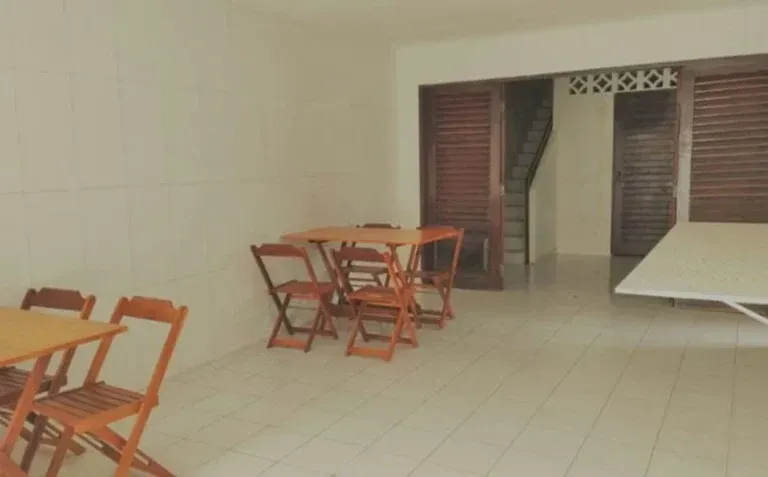 Sala vazia com azulejos, cadeiras e mesas de madeira, porta aberta com venezianas e um banco coberto à direita.