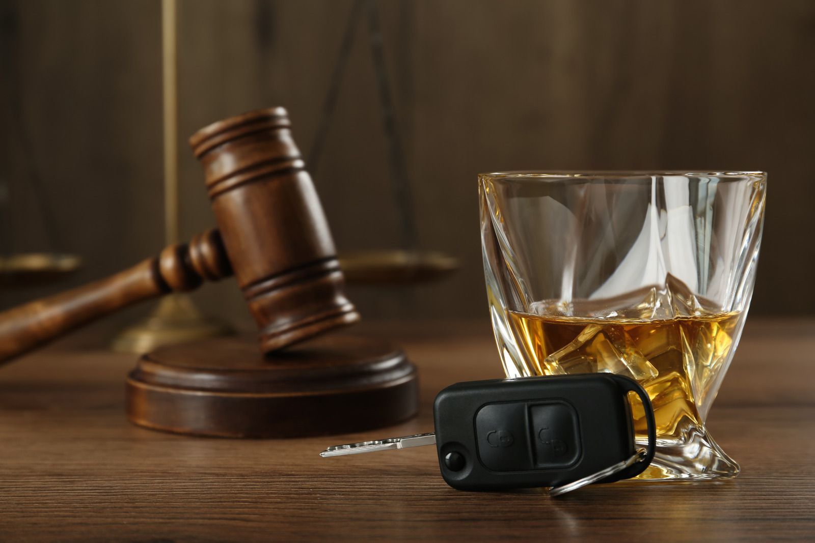 A glass of whiskey , a car key , and a gavel on a wooden table.