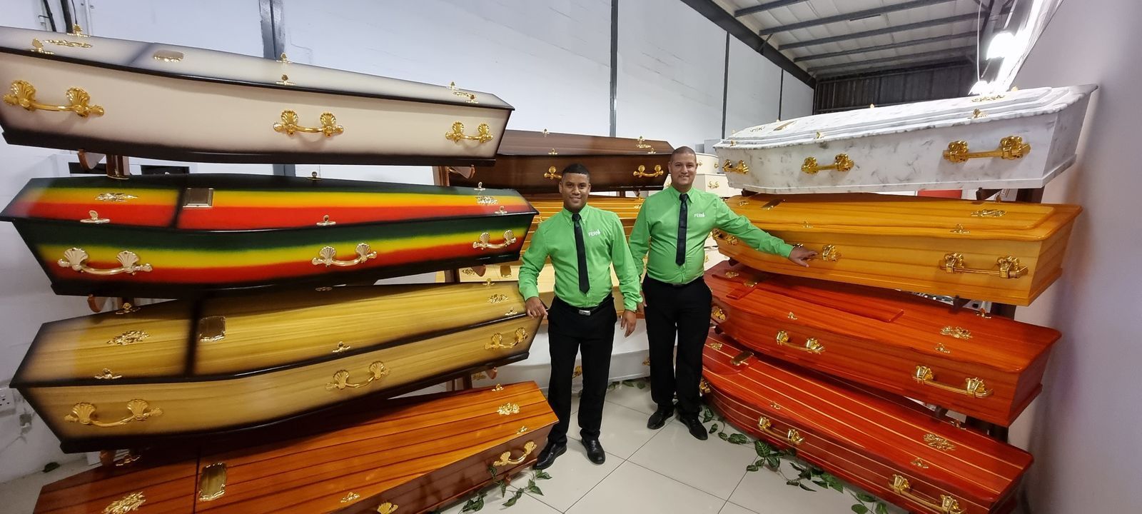Two people in green jackets standing beside stacks of colorful kayaks in a warehouse
