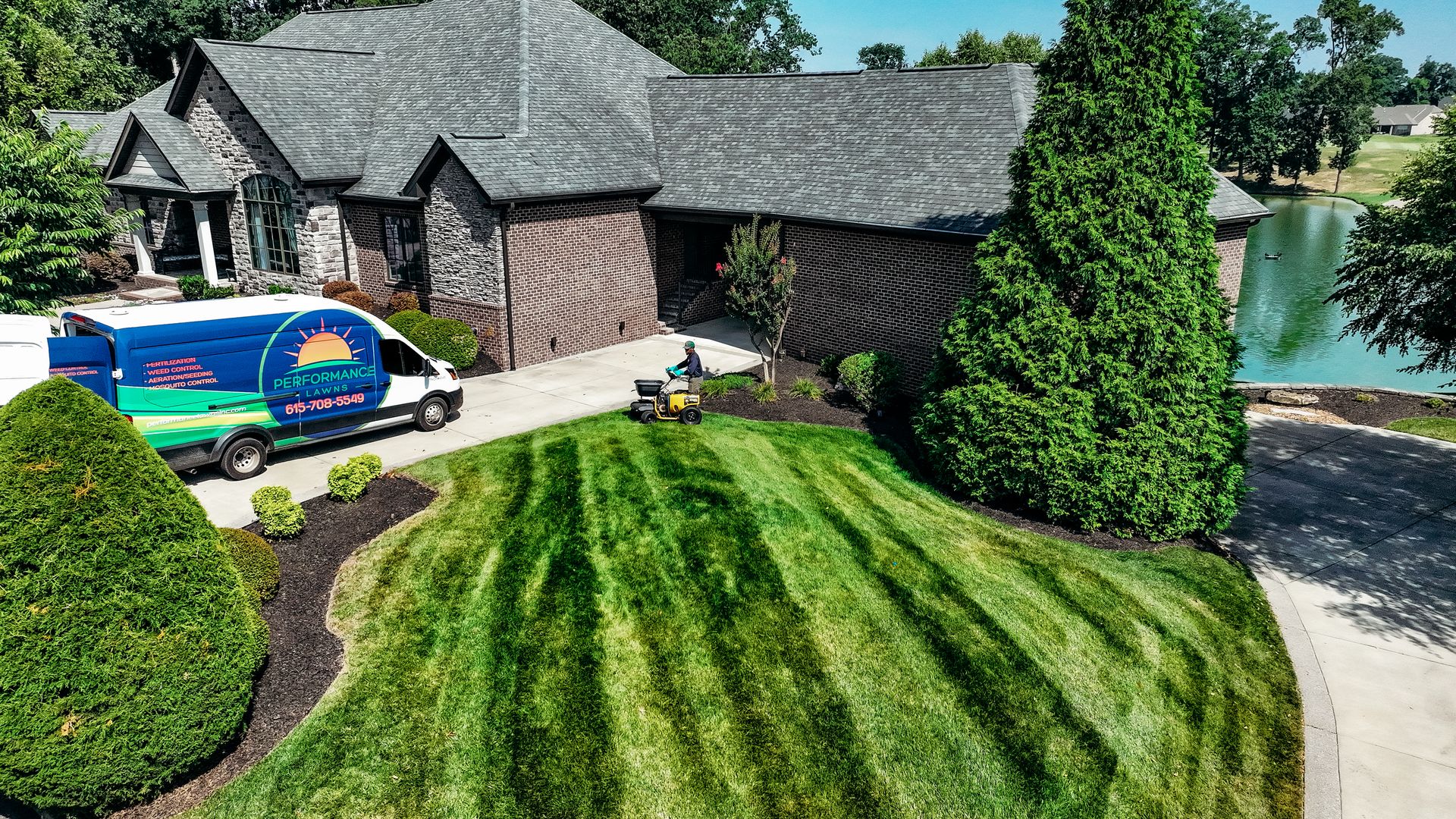 Lawn care service van parked in front of a large house as a worker mows the striped lawn near a pond.