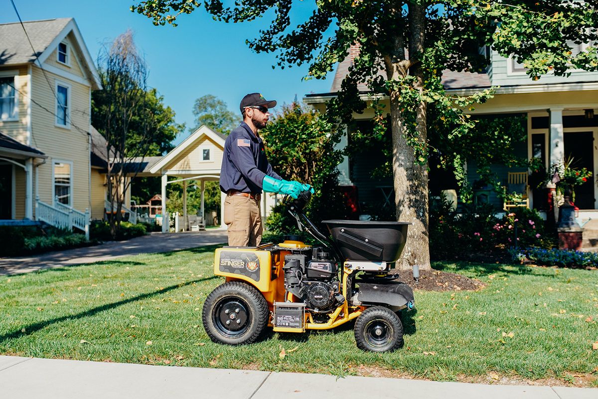 Man using a yellow lawn spreader in front of houses on a sunny day.