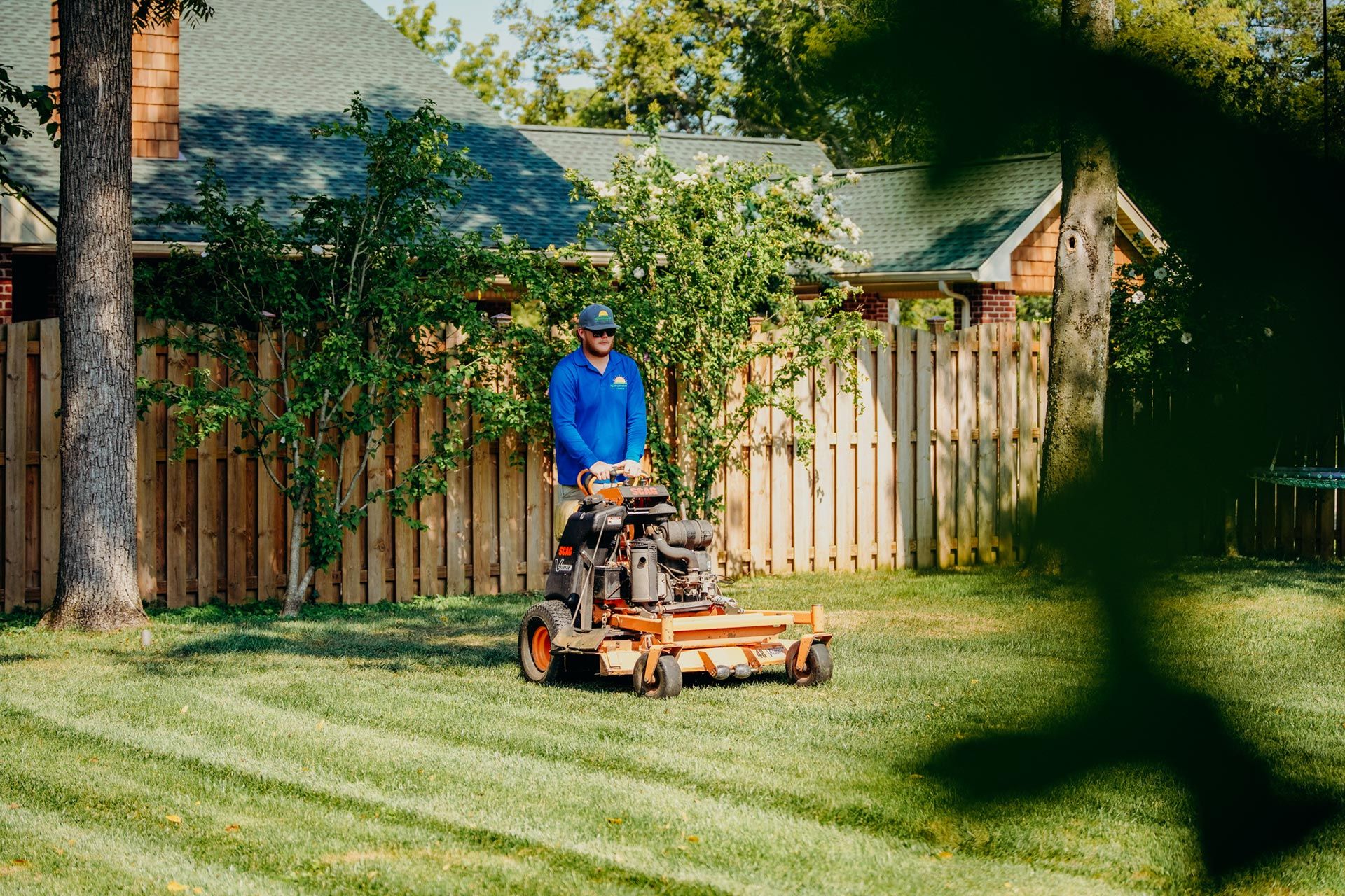 Man mowing a lawn with a riding mower in a backyard. Sunlight, trees, and a wooden fence are visible.