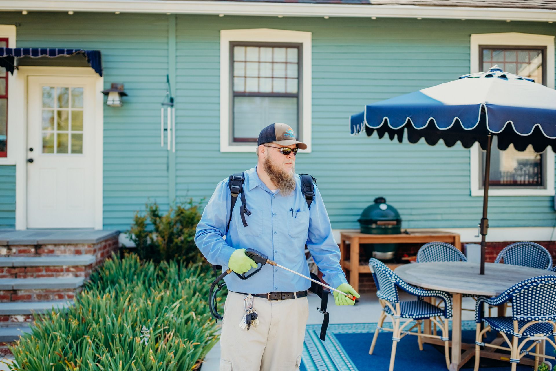 Man in work clothes spraying pesticide in a backyard; green house and patio furniture visible.