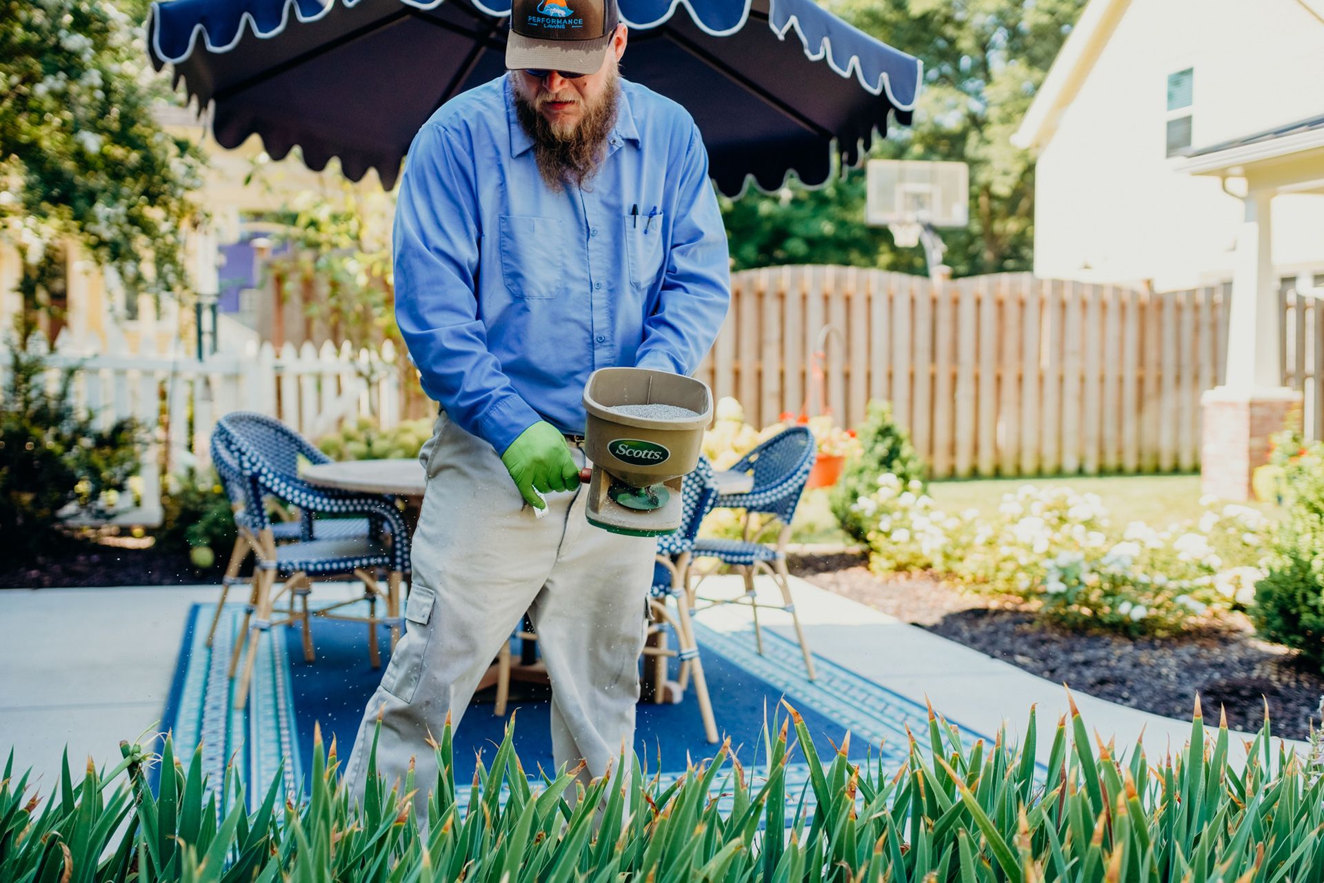 Man spreading fertilizer on lawn with a spreader in a backyard, under an umbrella.