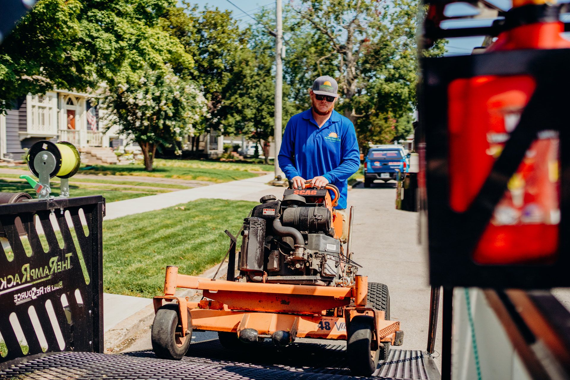 Man in blue pushes orange lawnmower onto a trailer on a sunny street.