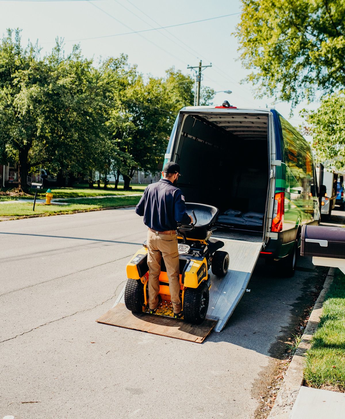 Man loading a yellow riding lawnmower into a truck on a residential street.