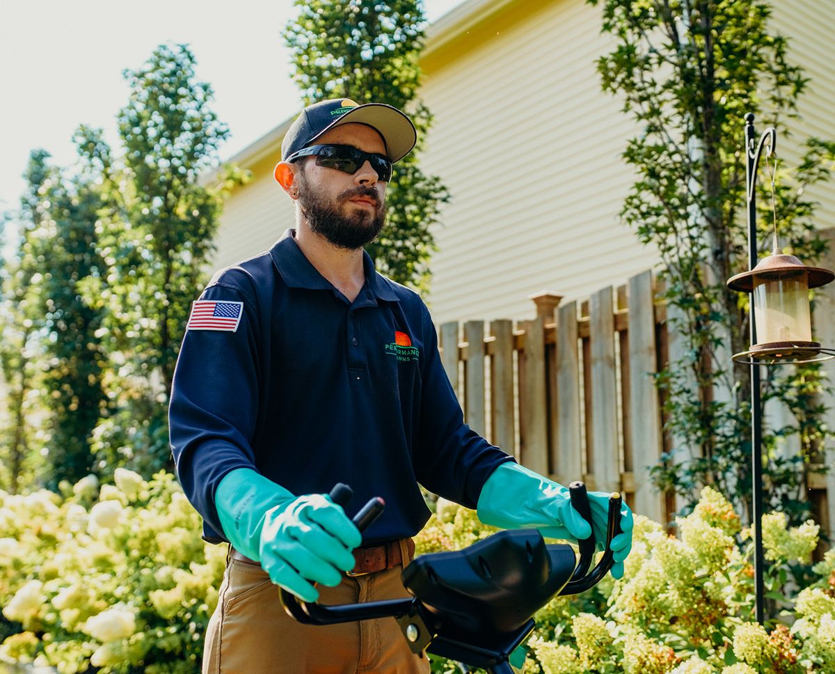 Man in uniform, gloves, and sunglasses operating equipment outdoors near a house and bushes.