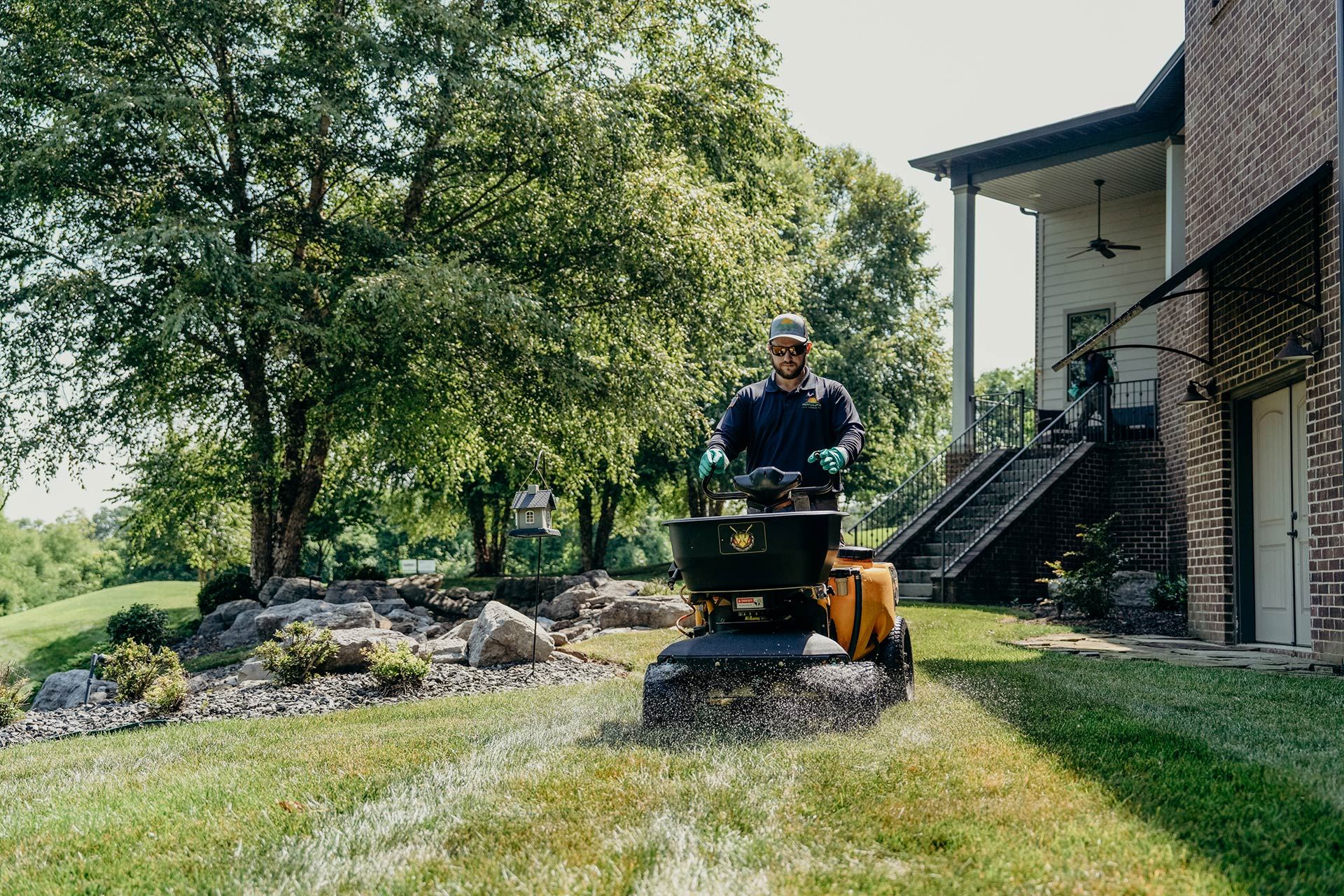 Man operating lawn spreader on green grass near a brick house and a rock garden.