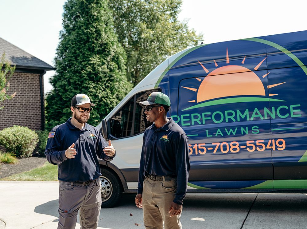 Two men in work attire stand near a Performance Lawns van. One gives a thumbs up.