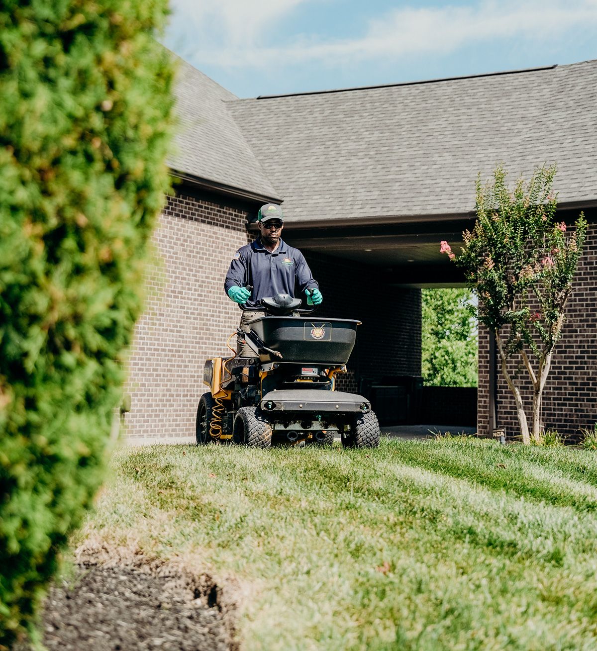 Man operating a lawn spreader on grass near a brick house.