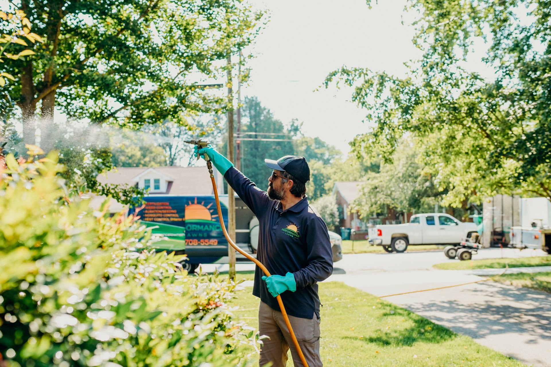 Man spraying a bush with a hose on a sunny day; green truck and houses in the background.